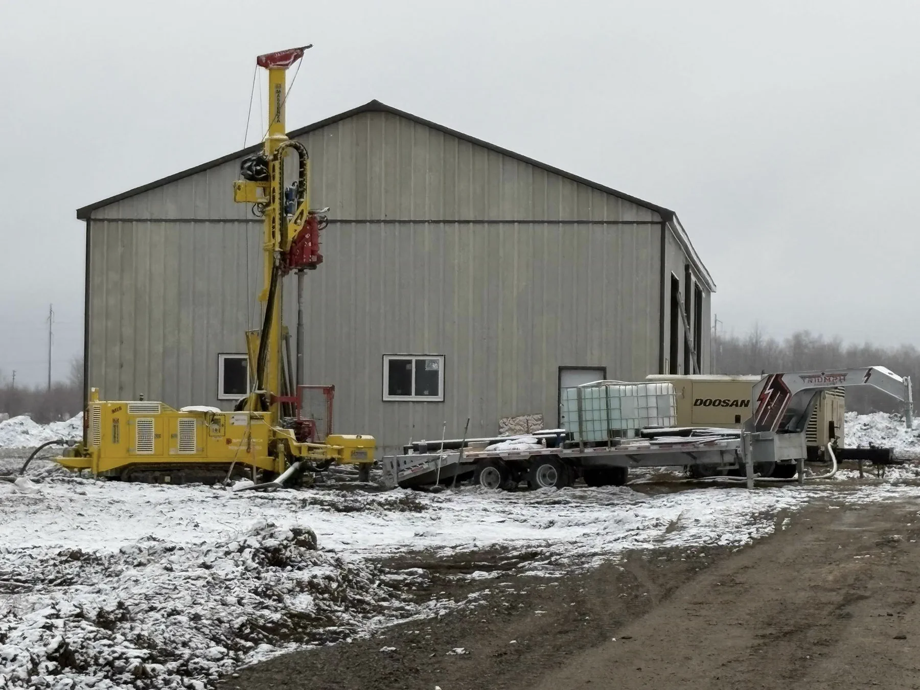 Tracked drill rig working near a shop building in snowy field conditions.